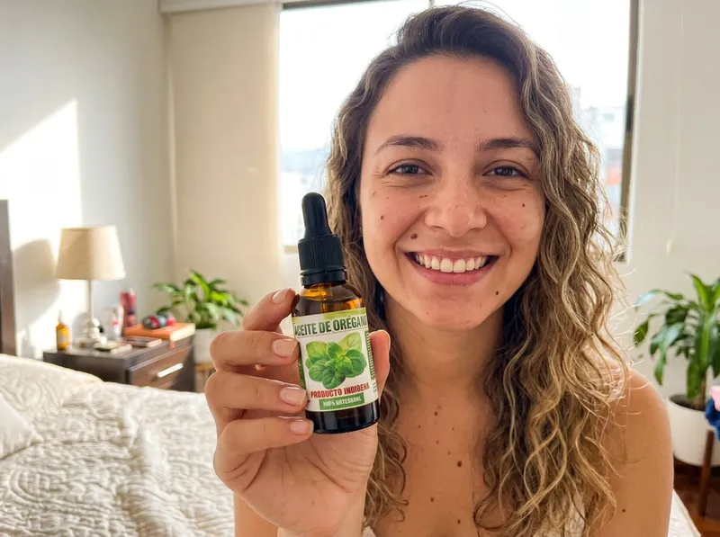 Una mujer colombiana de unos 27 años en su habitación iluminada en Cali, sosteniendo con confianza el frasco de aceite de orégano, foto natural de celular.